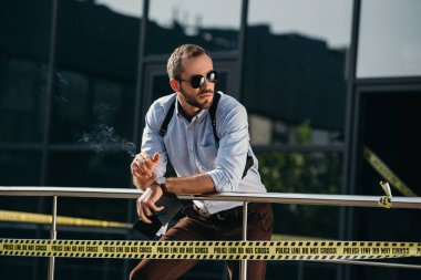 Sigara ve çit yaslanmış yan isteyen güneş gözlüklü erkek dedektif