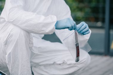 cropped view of criminologist in protective suit and latex gloves taking evidence into flask at crime scene clipart