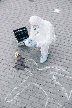 male criminologist in protective suit and latex gloves looking at chalk line with blood spot at crime scene clipart