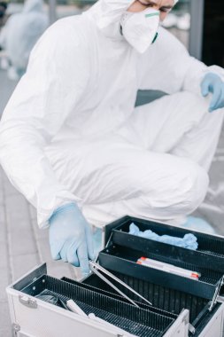 male criminologist in protective suit and latex gloves taking tweezers from investigation tools bag  clipart