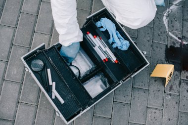 partial view of criminologist in protective suit  sitting near investigation tools clipart