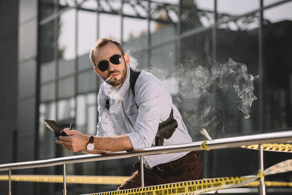 мужчина-детектив курит и делает заметки, опираясь на забор
