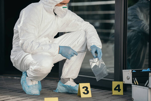 male criminologist in protective suit and latex gloves packing evidence at crime scene