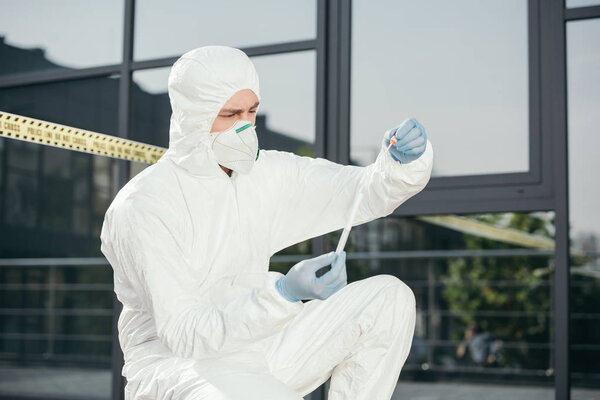 male criminologist in protective suit and latex gloves looking at evidence at crime scene