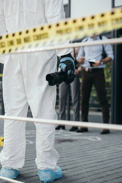 cropped view of criminologist in protective suit and latex gloves with camera at crime scene