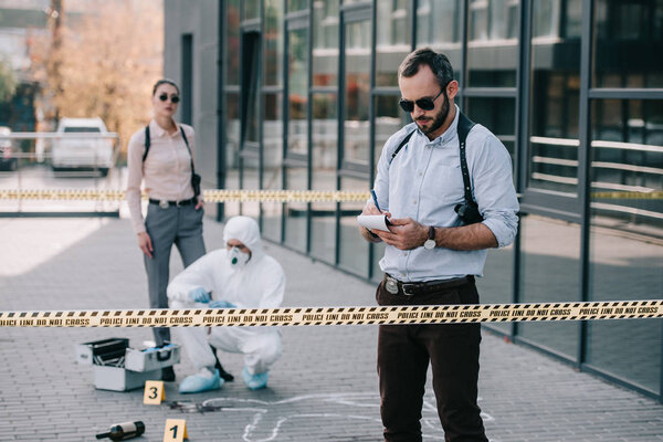 Детектив-мужчина, стоящий на месте преступления и записывающий в записную книжку
