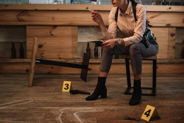 裁剪视图吸烟女侦探坐在犯罪现场与证据标记和尸体轮廓 — 图库照片