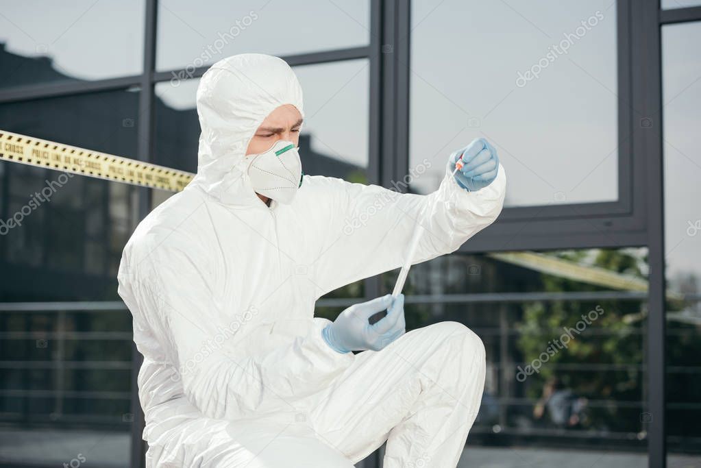 male criminologist in protective suit and latex gloves looking at evidence at crime scene