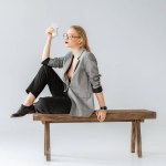 Stylish girl holding glass of milk and sitting on wooden bench on grey