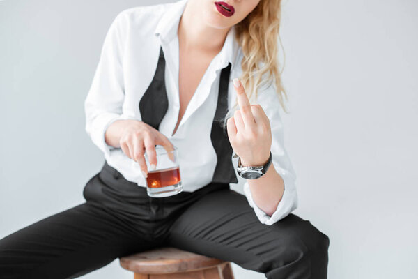 обрезанный вид элегантной женщины со стаканом виски, сидящего на стуле и показывающего средний палец, изолированный на сером
