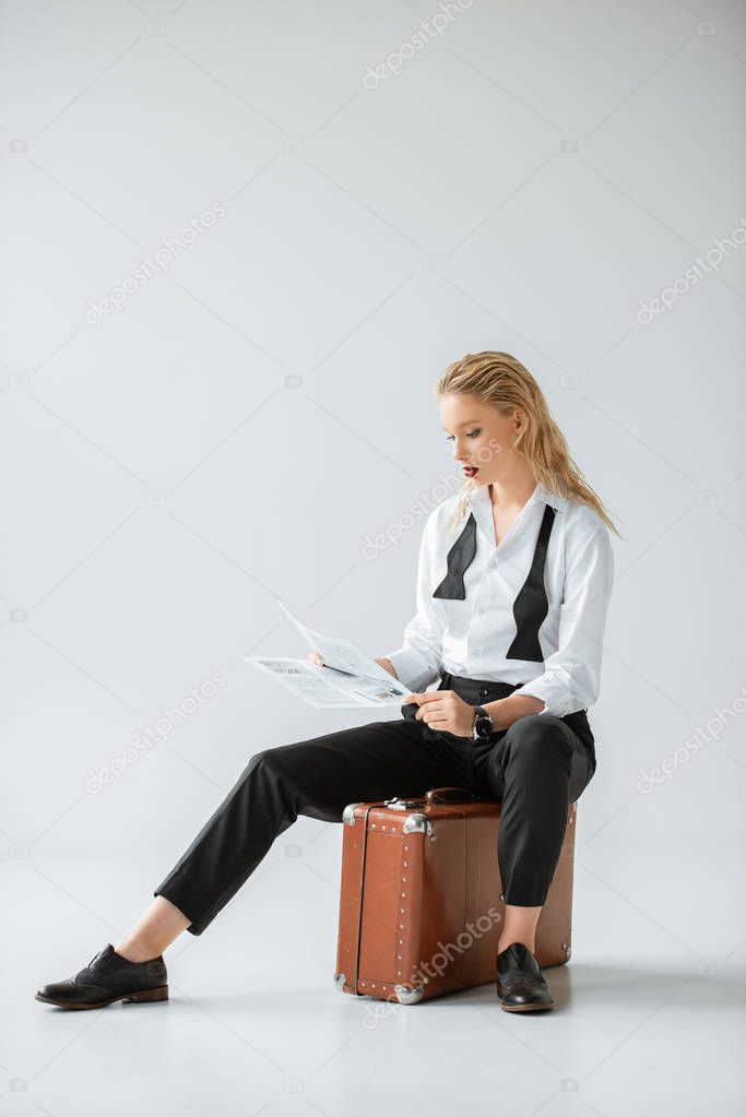 fashionable girl reading newspaper while sitting on retro suitcase on grey