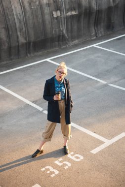 stylish young woman with coffee to go posing on parking clipart