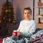 Young blonde woman sitting on couch, holding cup of hot cocoa with marshmallows and looking at camera at christmas time