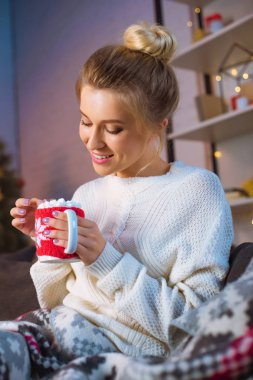smiling young blonde woman covered in blanket sitting on couch and holding cup of hot cocoa with marshmallows at christmas time  clipart