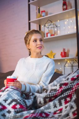 young blonde woman covered in blanket sitting on couch and holding cup of hot cocoa with marshmallows at christmas time  clipart