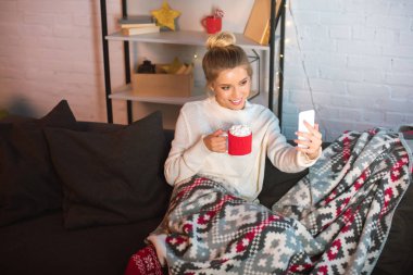 mutlu sarışın genç kadın kanepede oturan, Marshmallow ile sıcak kakao fincan holding ve selfie smartphone üzerinde Noel zamanda alarak battaniyeye örtülü 