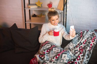 young happy blonde woman covered in blanket sitting on couch, holding cup of hot cocoa with marshmallows and taking selfie on smartphone at christmas time  clipart