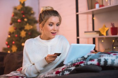kredi kartı tutan ve online alışveriş için Noel zamanda dijital tablet kullanarak kanepede oturan sürpriz genç sarışın kadın