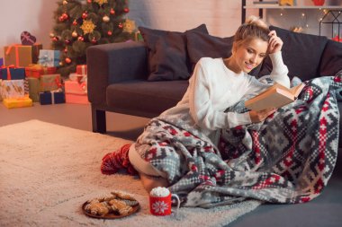 Charming girl smiling and reading book at home on Christmas eve clipart