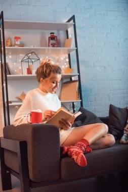 Sarışın genç kadın fincan sıcak kakao ve kanepe ve okuma kitabı Noel zamanda oturma marshmallows ile gülümseyen 
