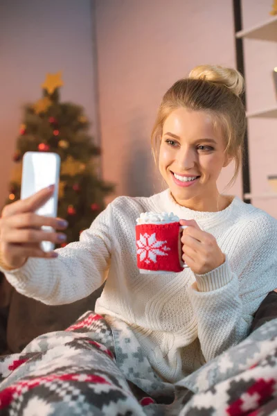 愉快的年轻金发碧眼的女人拿着一杯热可可与棉花糖和采取自拍在智能手机在圣诞节时间 — 图库照片