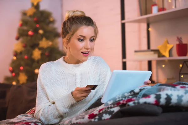 惊讶年轻的金发碧眼的女人坐在沙发上拿着信用卡 并在圣诞节时间使用数字平板电脑在线购物 — 图库照片