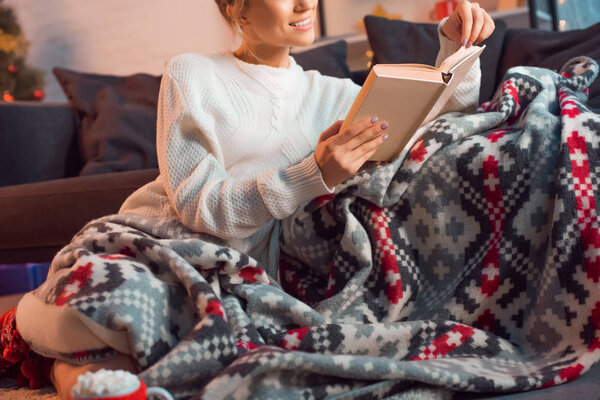 красивая улыбающаяся женщина читает книгу дома в узорчатом одеяле в канун Рождества

