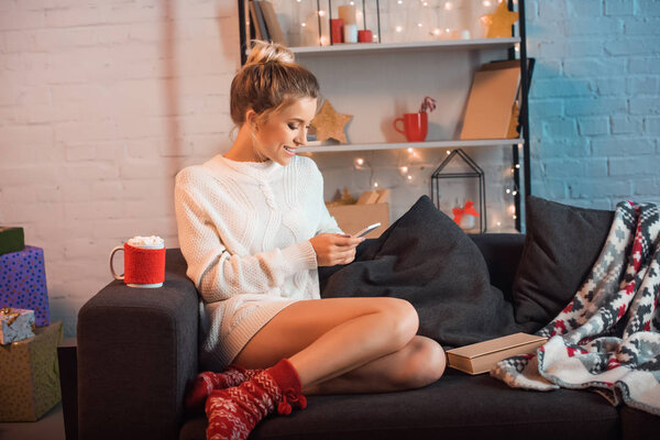 smiling young blonde woman sitting on couch and using smartphone at christmas time 