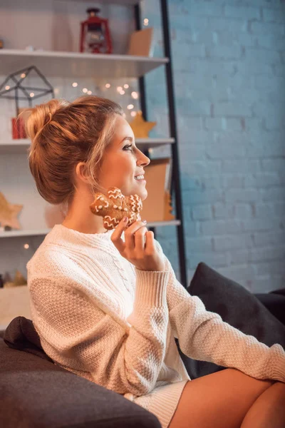 微笑的年轻金发碧眼的女人坐在沙发上 摆姿势 拿着姜饼饼干在圣诞节时间 — 图库照片