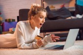 überrascht junge blonde Frau am Boden liegend, mit Kreditkarte, Laptop und Online-Shopping zur Weihnachtszeit