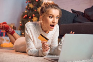 Genç Sarışın kadın yerde yatarken, kredi kartı holding, dizüstü bilgisayar kullanarak ve Noel zamanda online alışveriş yaparken heyecanlı 