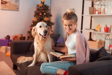 Noel zamanda golden retriever köpek ve okuma kitap ile kanepede oturan güzel sarışın genç kadın