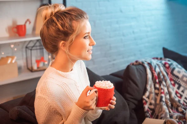 ソファに座っているとクリスマスの時にマシュマロとココアのカップを保持している美しい若い金髪女性 — ストック写真