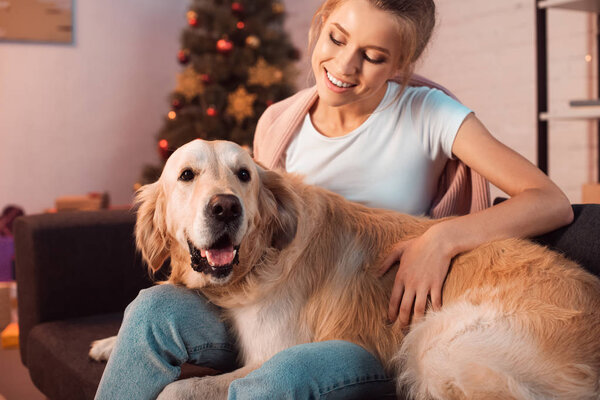 beautiful smiling young blonde woman sitting on couch and hugging golden retriever dog at christmas time