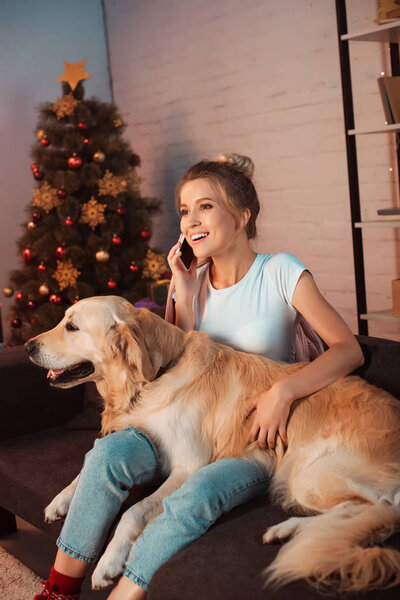 beautiful smiling young blonde woman sitting on couch with golden retriever dog and talking on smartphone at christmas time