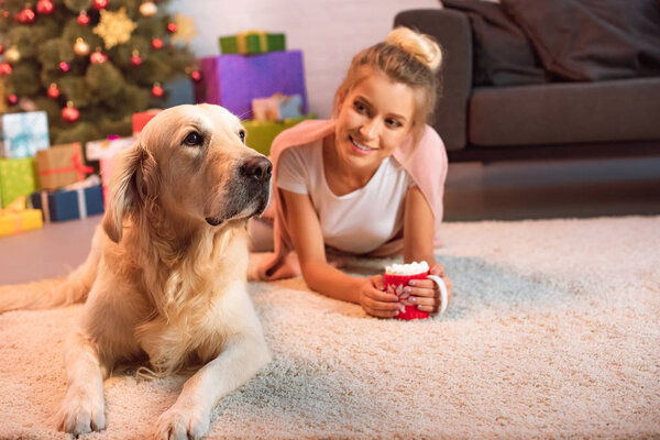 beautiful young blonde woman lying on fluffy rug with golden retriever dog and cup of hot cocoa with marshmallows at christmas time 