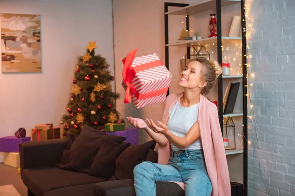 欢快的年轻金发碧眼的女人坐在沙发上 并在圣诞节时间在空气中抛出礼物 — 图库照片
