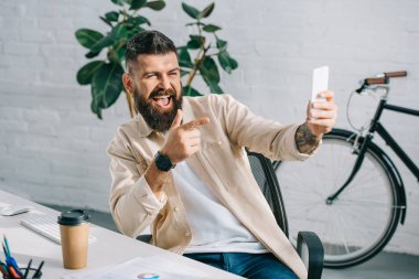 Bearded businessman pointing at camera while taking selfie in office chair clipart