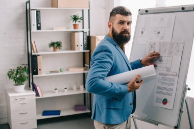 handsome adult male architect in blue formal wear holding blueprint, using flip chart and working on project in office clipart