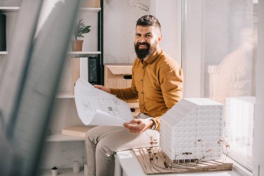 gülümseyen blueprint tutarak ve Office proje üzerinde ev modeli oturan yetişkin erkek mimar sakallı 