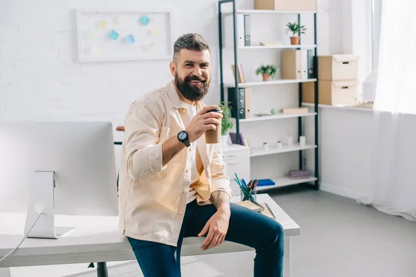 Smiling bearded businessman sitting on office desk with paper cup