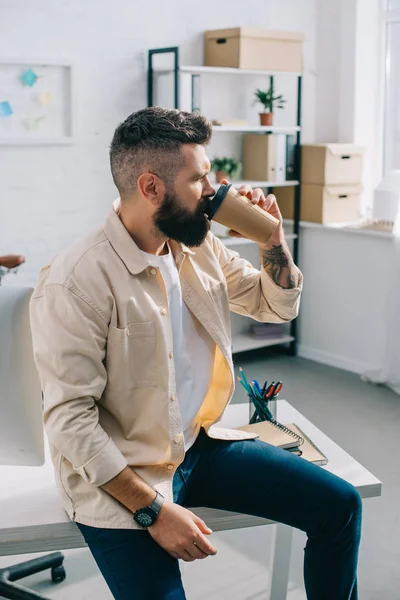 ひげを生やしたビジネスマンの机の上に座っていると オフィスで紙コップのコーヒーを飲む  — 無料ストックフォト