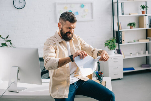 Aggressive boss sitting at desk and tearing papers in modern office 