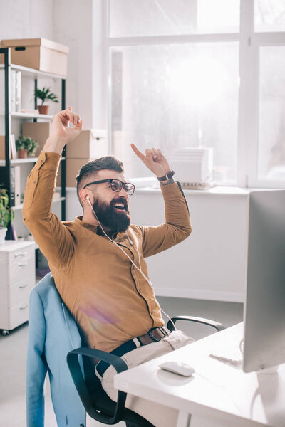 happy bearded adult businessman in earphones sitting at computer and listening to music desk in office