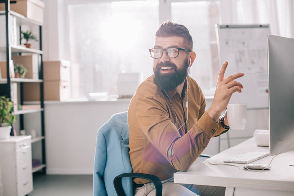 smiling bearded adult businessman in earphones sitting and working at computer desk in office