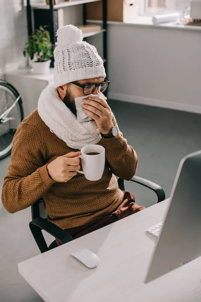生病的商人戴着围巾和针织帽子坐在办公桌前 拿着一杯茶和打喷嚏 — 免费的图库照片