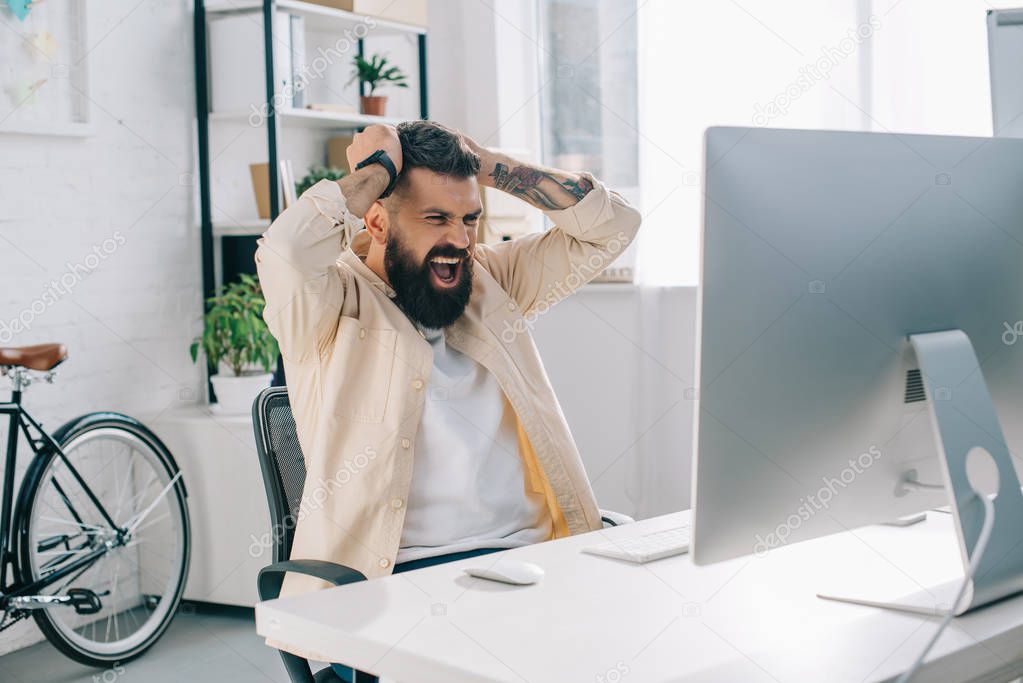 Impulsive bearded businessman with hands on head sitting at computer desk and screaming in office