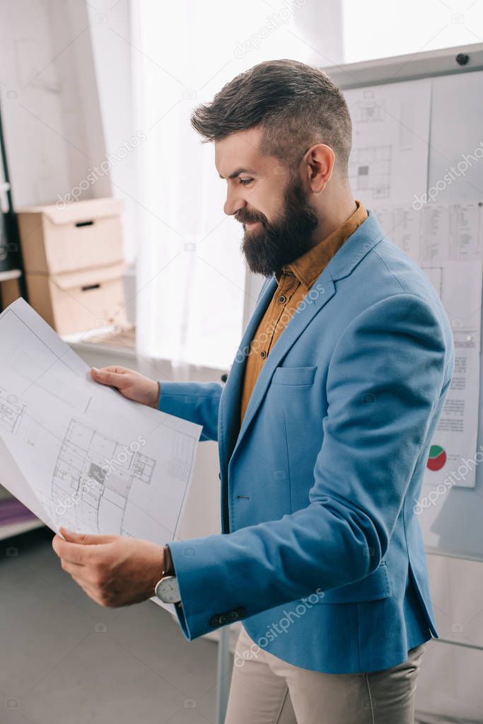 side view of smiling adult male architect in blue formal wear holding blueprint, using flip chart and working on project in office