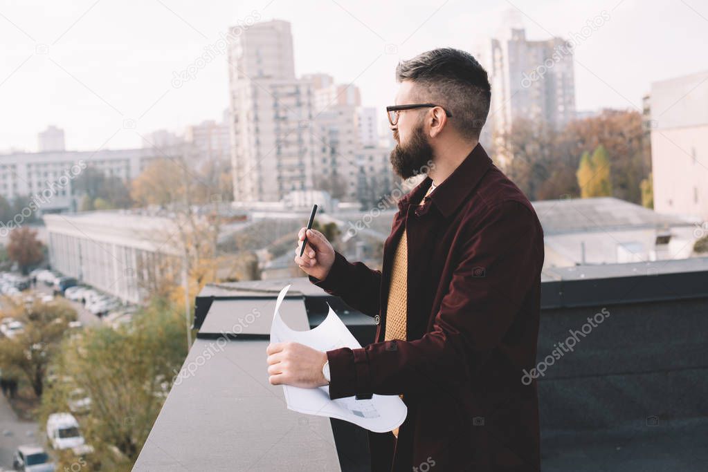 adult male architect in glasses holding blueprint and working on rooftop with beautiful view