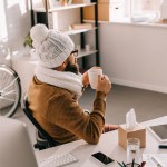 Sick businessman in scarf and knitted hat sitting at office desk and holding cup of tea